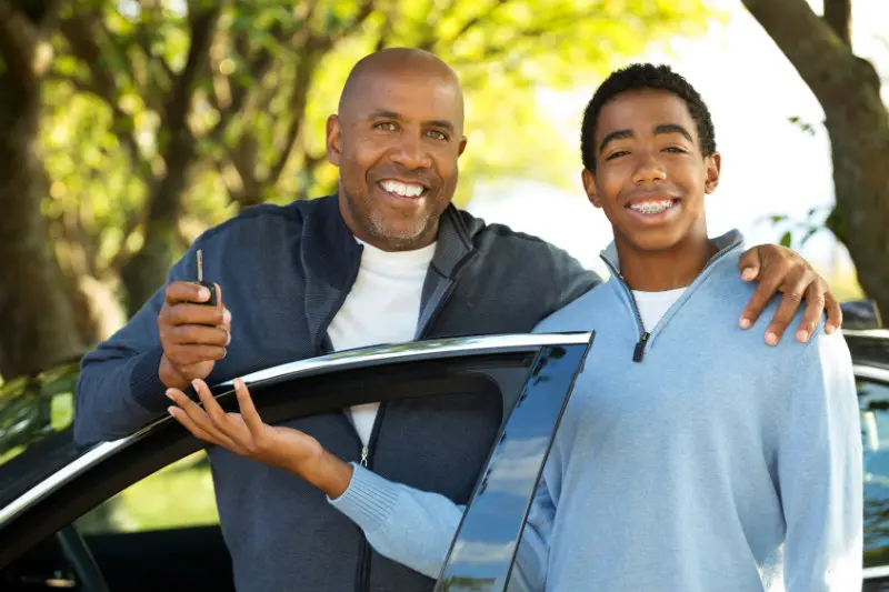 A smiling father and son standing in front of their car.