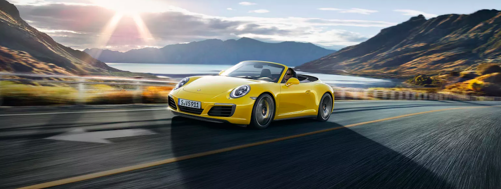 A yellow sports car driving down the road with mountains and a lake in the back
